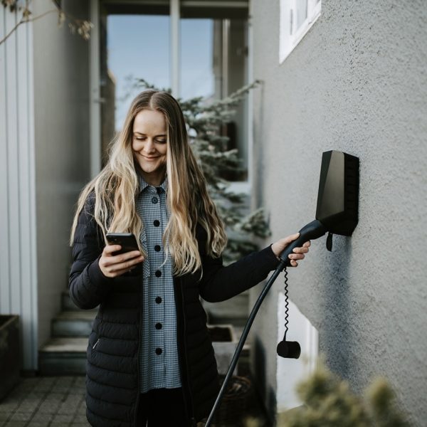 Ung kvinne med langt lyst hår styrer sort elbillader på grå murvegg med en smarttelefon